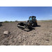 Front view of tracked skid steer with open rock reel attachment working on cleared soil under clear sky