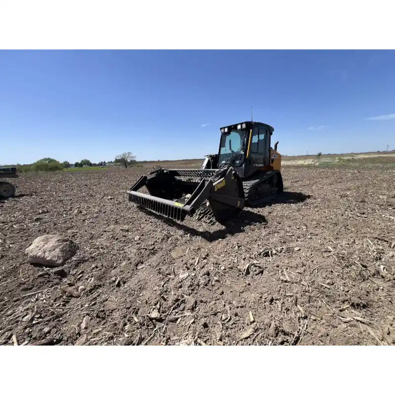 Front view of tracked skid steer with open rock reel attachment working on cleared soil under clear sky