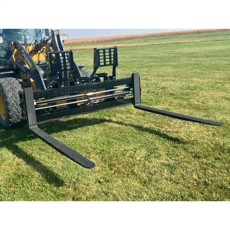 Black steel pallet fork attachment with adjustable tines mounted on a yellow skid steer on grassy field