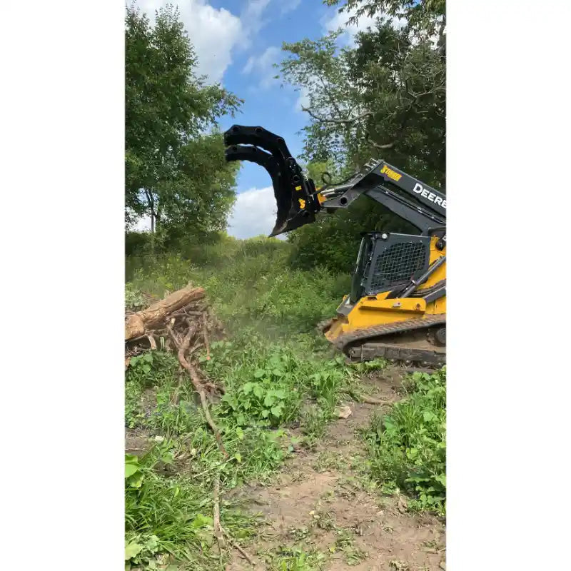 Yellow John Deere skid steer with black claw-style grapple attachment lifting tree roots in a grassy, wooded area