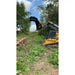 Yellow Deere skid steer with black clam-style grapple attachment raised above uprooted tree and green foliage under blue sky