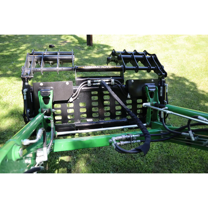 Close-up of black steel grapple attachment with hydraulic hoses mounted on green compact tractor on grass outdoors
