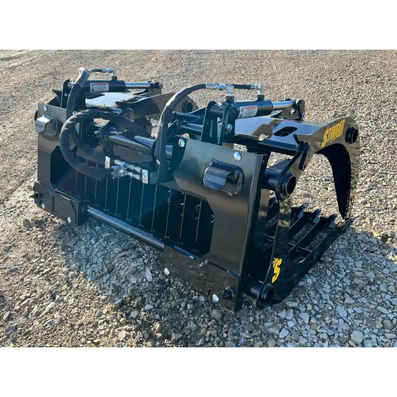 Black steel rock grapple attachment with hydraulic arms and wide spaced tines resting on gravel ground
