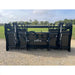 Wide black steel clam grapple attachment with hydraulic cylinders and hoses resting on gravel outdoors
