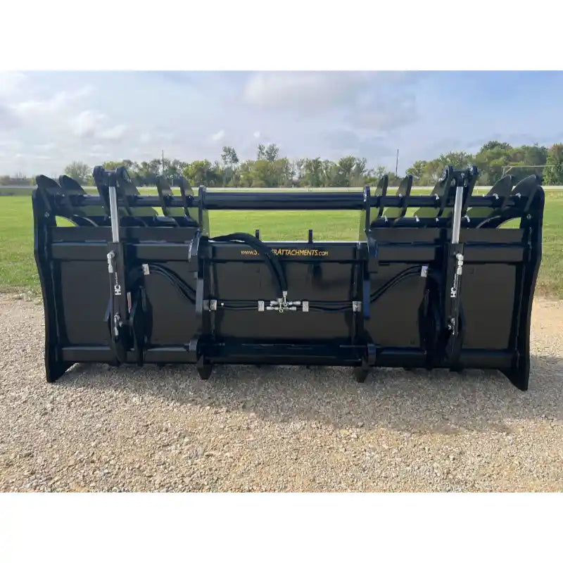 Wide black steel clam grapple attachment with hydraulic cylinders and hoses resting on gravel outdoors