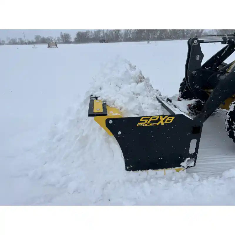 Yellow and black skid steer snow pusher attachment angled left, pushing snow on flat, open snow-covered field outdoors