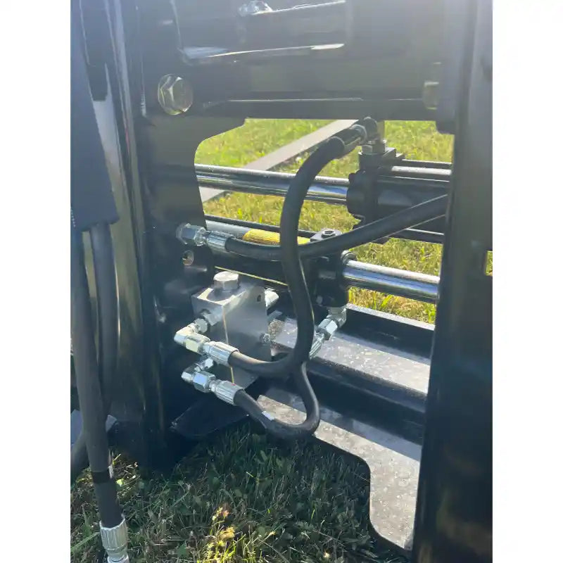 Close-up of hydraulic hose connections and metal fittings on black skid steer pallet fork attachment over grass field