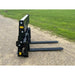 Black steel skid steer pallet forks with perforated vertical backrest and two long flat tines on gravel surface