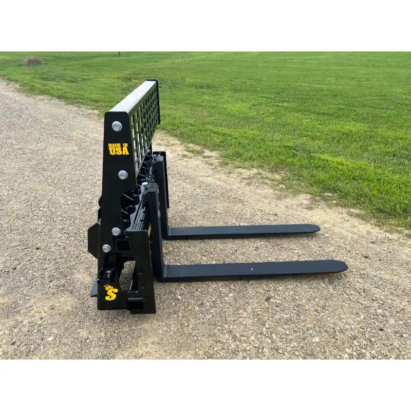 Black steel skid steer pallet forks with perforated vertical backrest and two long flat tines on gravel surface