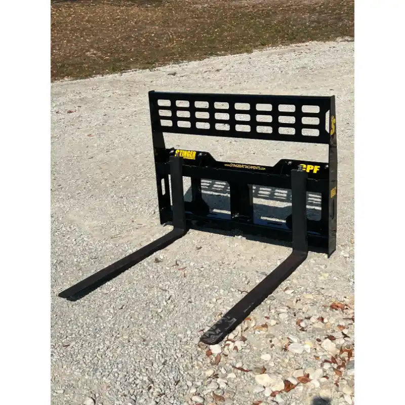 Black steel forklift pallet fork attachment with rectangular perforated backrest and two extended tines on gravel ground