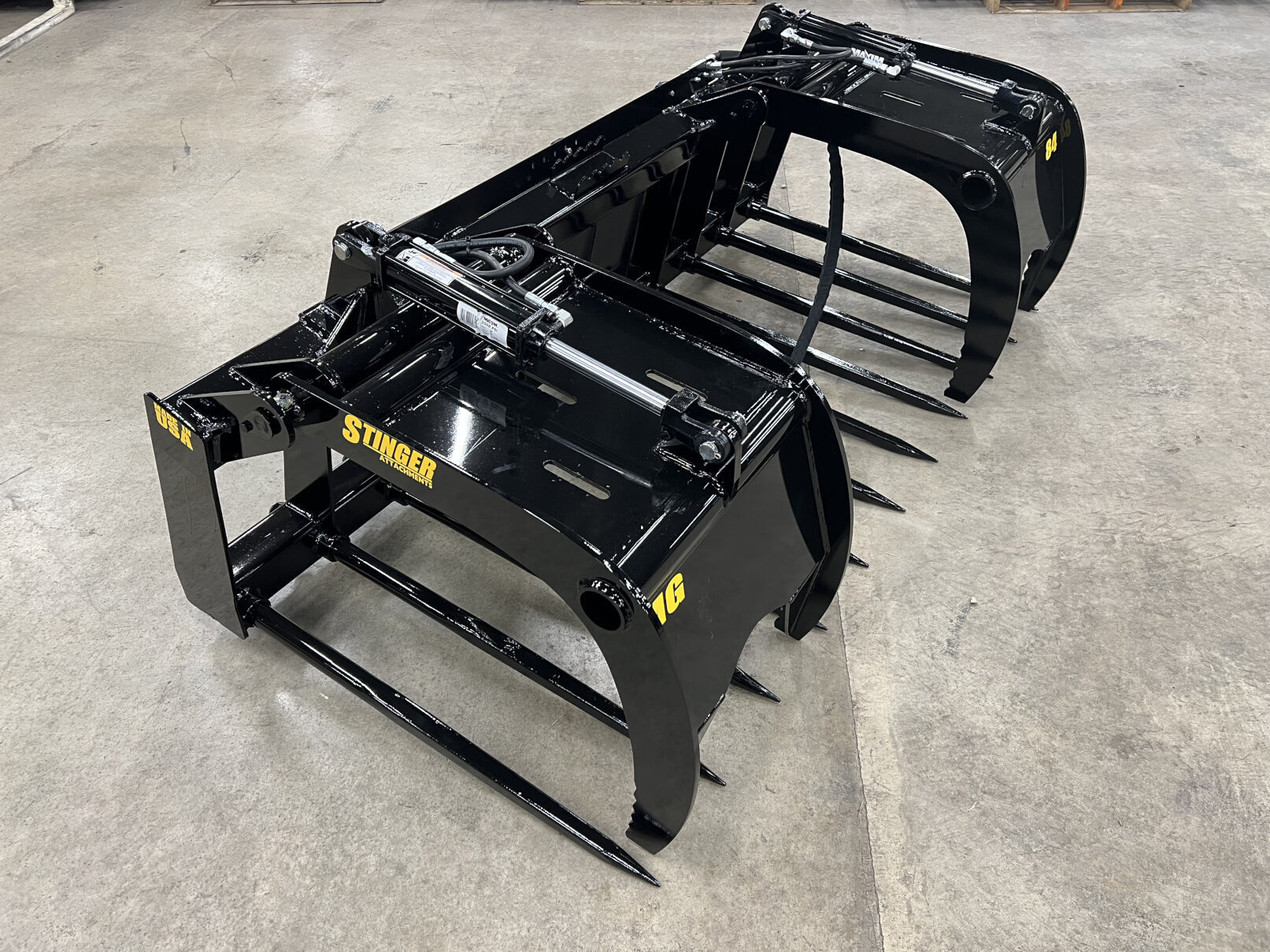 Pair of black hydraulic grapple attachments with teeth, resting on concrete floor indoors