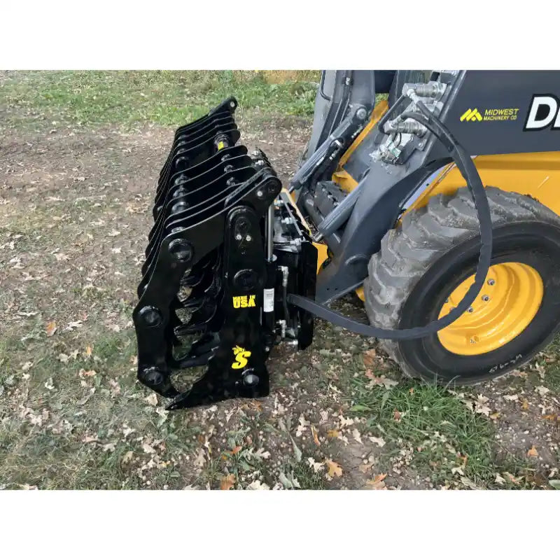 Black steel clam grapple attachment connected to skid steer loader with hydraulic hoses on grassy ground outdoors