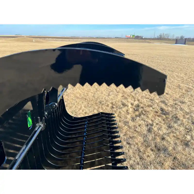 Close-up of black metal grapple attachment with serrated curved upper jaw and spaced tines over dry grass field