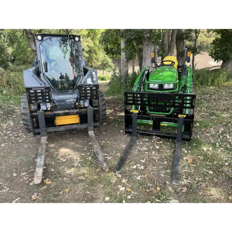 Two black steel skid steer and tractor pallet fork attachments with grid backrests and extended forks on grass and dirt outdoors
