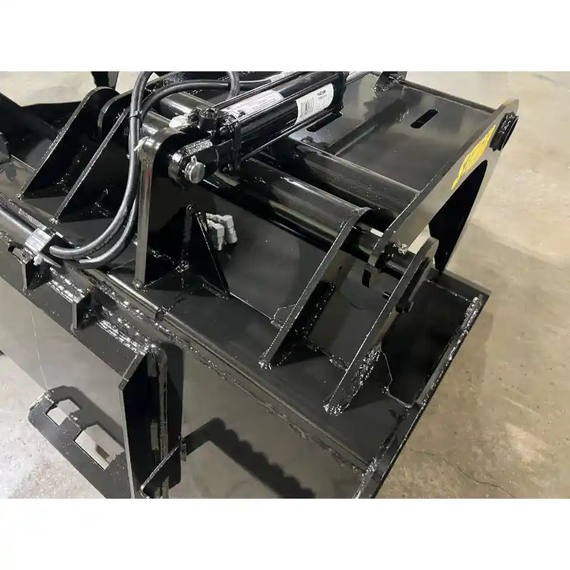 Black steel demolition grapple bucket attachment showing hydraulic cylinder, hoses, and reinforced welds on indoor concrete floor