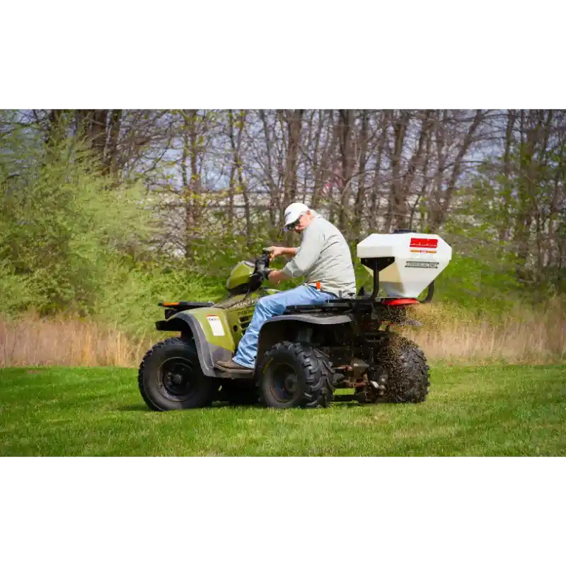 Operator riding ATV fitted with Swisher spreader dispersing material over grass from rear, gallery position 1