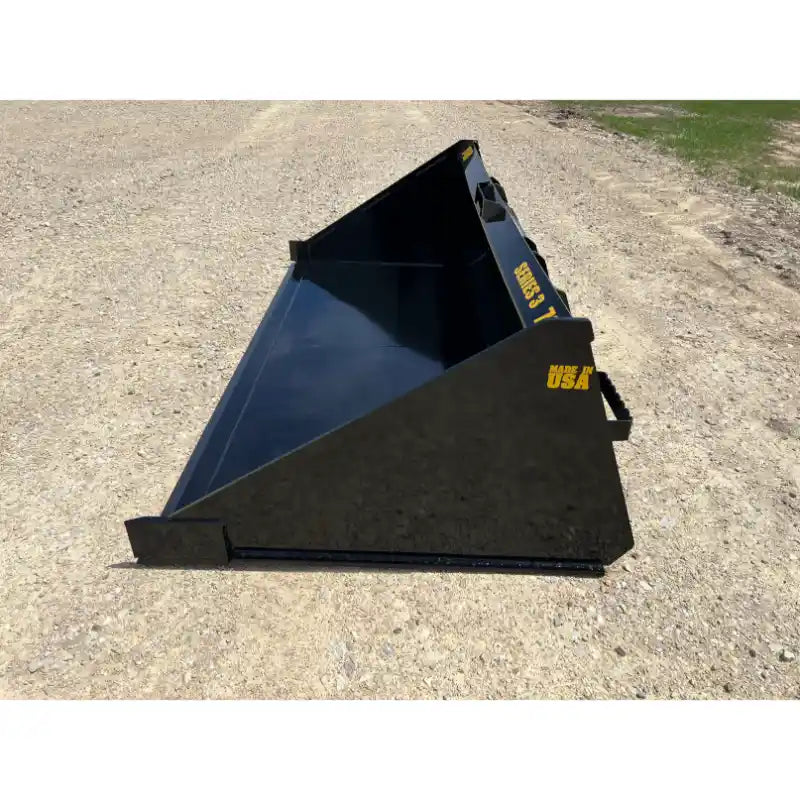 Black skid steer material bucket with smooth interior and angled sides resting on gravel surface