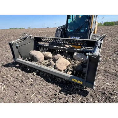 Low-angle front view of skid steer with rock bucket attachment holding large rocks on tilled soil under clear sky