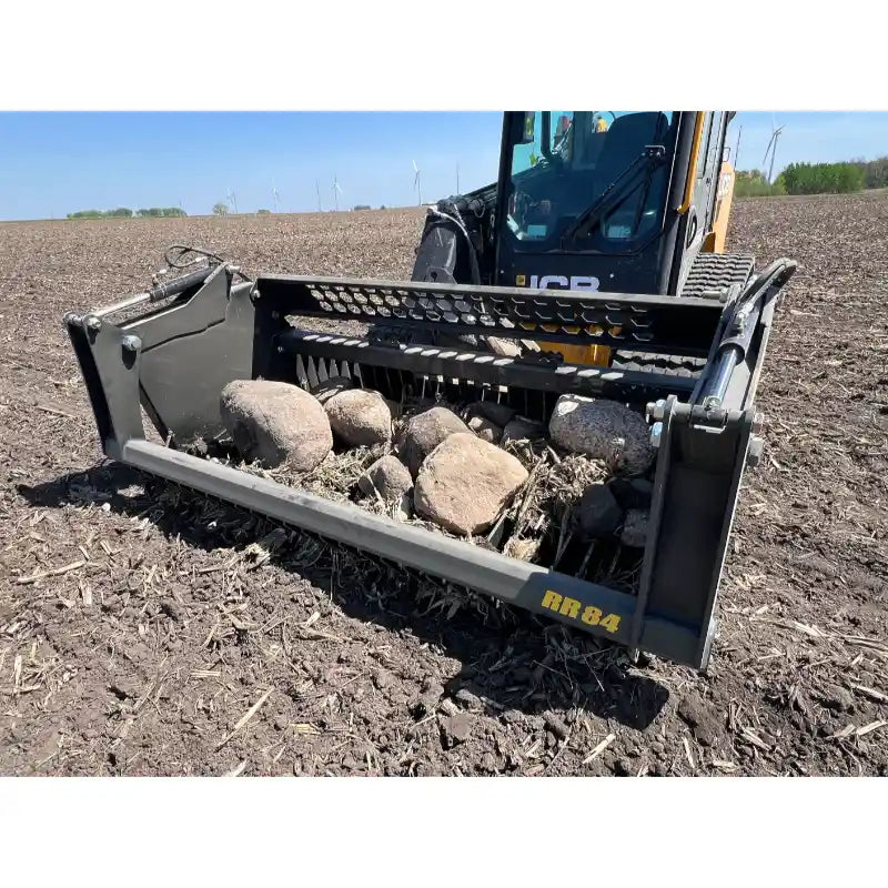 Close-up view of skid steer with metal rock bucket reel attachment holding large rocks and debris on tilled soil under clear sky