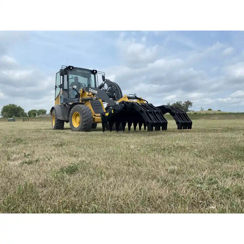 Yellow Deere loader equipped with black steel clam-style grapple attachment lowered over dry grassy field under cloudy sky