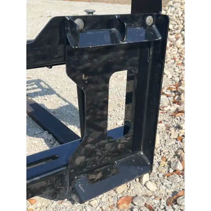 Close-up of black steel skid steer pallet fork mounting bracket with rectangular cutout on gravel surface outdoors