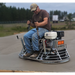 Operator steering a low rider ride-on power trowel smoothing a large freshly poured concrete slab outdoors