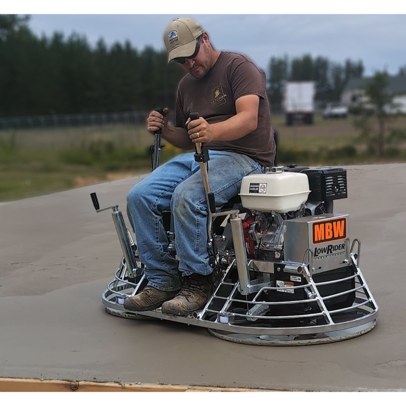 Operator steering a low rider ride-on power trowel smoothing a large freshly poured concrete slab outdoors