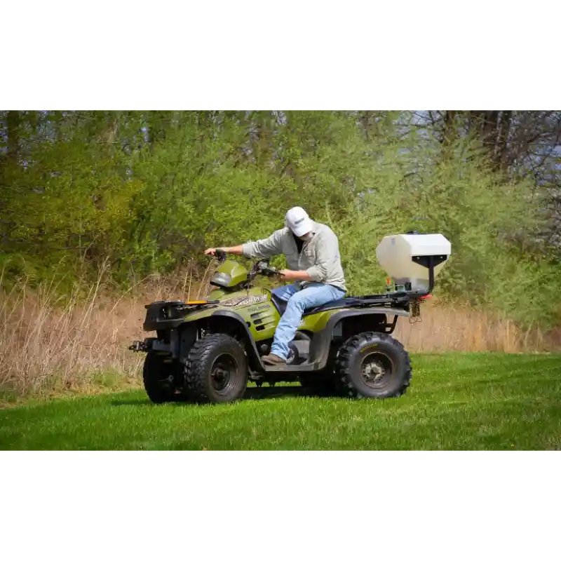 Operator on green ATV with white Swisher ATV Commercial Pro Spreader attached at rear on grass, gallery position 1
