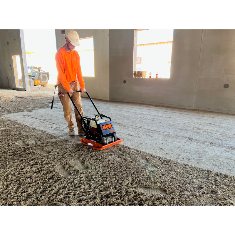 Construction worker in orange shirt and beige pants operating orange and black MBW plate compactor on gravel floor inside building