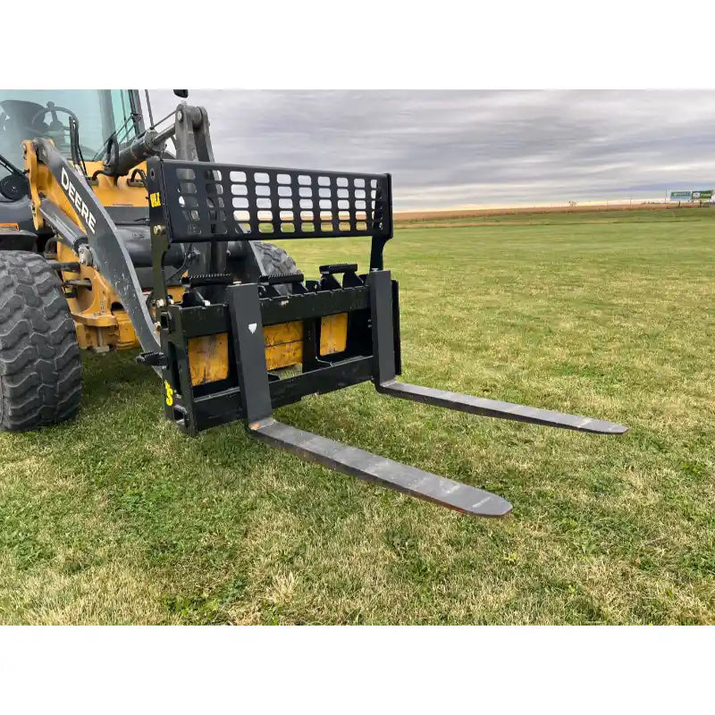 Yellow skid steer loader with black pallet fork attachment featuring two flat steel tines on grass field