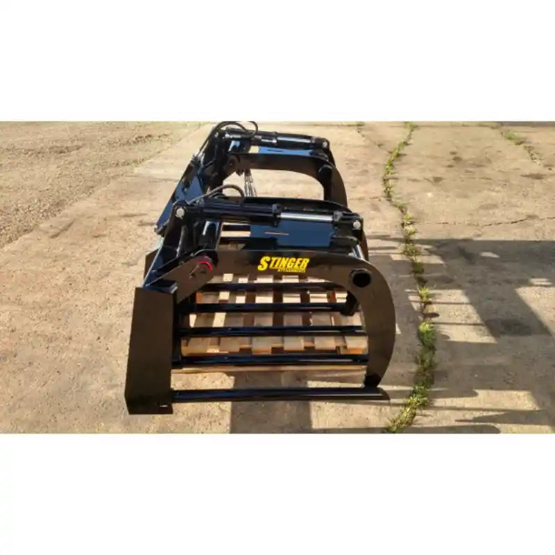 Black-painted steel manure tine grapple attachment with spaced horizontal tines and hydraulic linkage on concrete surface outdoors