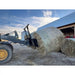 Skid steer attachment lifting a large round hay bale outdoors near a barn under partly cloudy sky