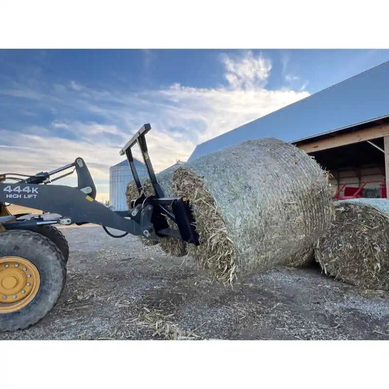 Skid steer attachment lifting a large round hay bale outdoors near a barn under partly cloudy sky