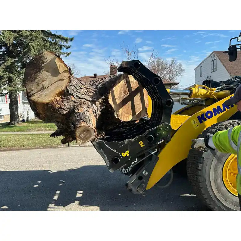 Yellow loader with black hydraulic clam-style grapple holding large cut tree trunk on paved surface