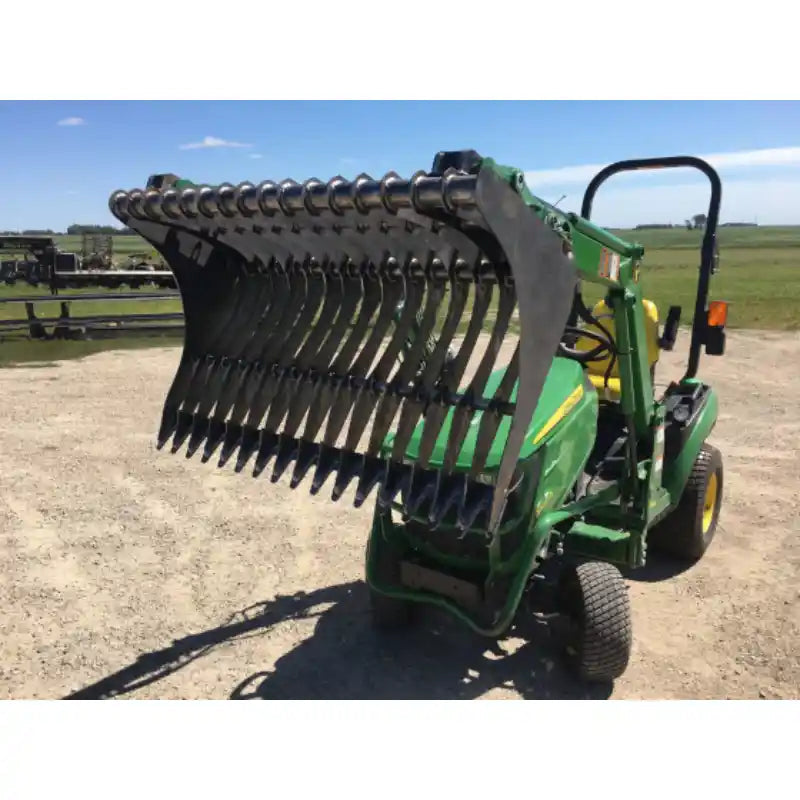 Green compact loader with a rock bucket attachment featuring closely spaced curved tines lifted above gravel ground