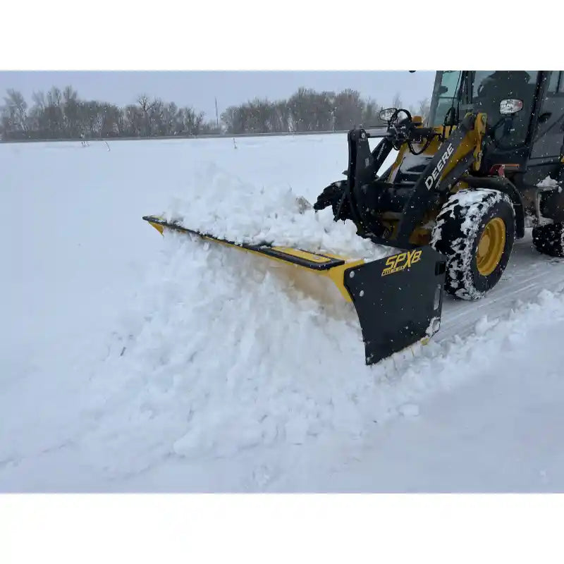 Yellow skid steer snow pusher attachment moving snow on a flat, snow-covered field with Deere loader wheels visible