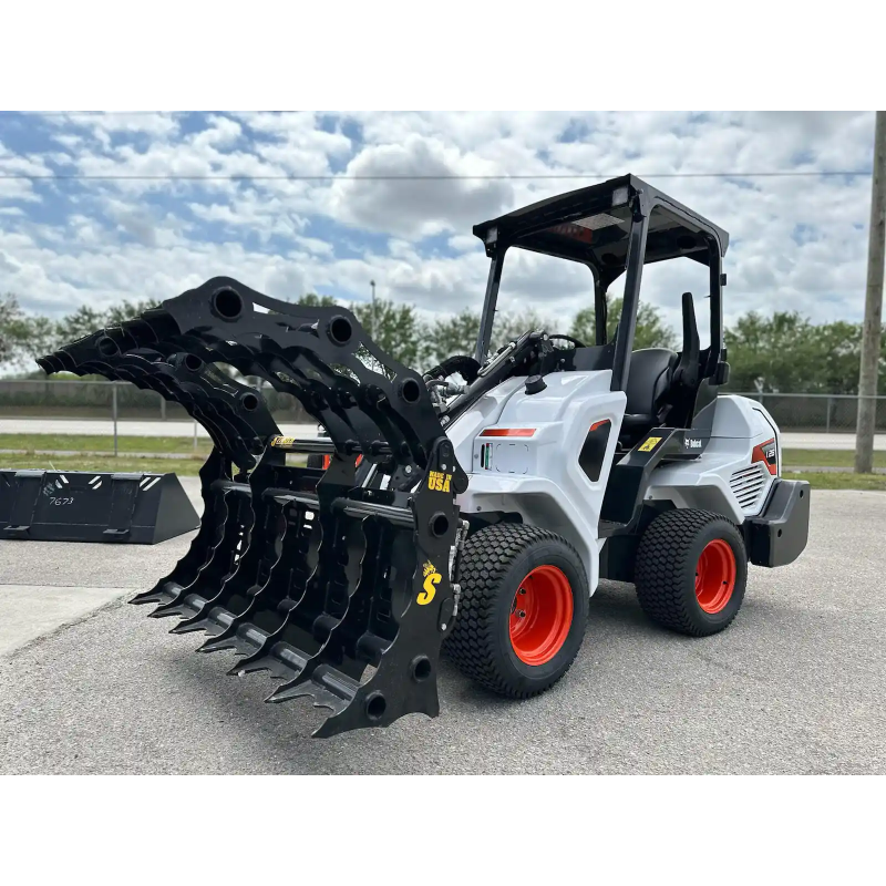 White skid steer with black multi-tine grapple attachment and orange wheels parked on asphalt near fence under cloudy sky