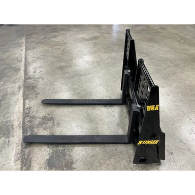 Black painted steel pallet fork attachment with two long horizontal tines and reinforced vertical mounting plate on concrete floor