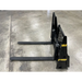 Black painted steel pallet fork attachment with two long horizontal tines and reinforced vertical mounting plate on concrete floor