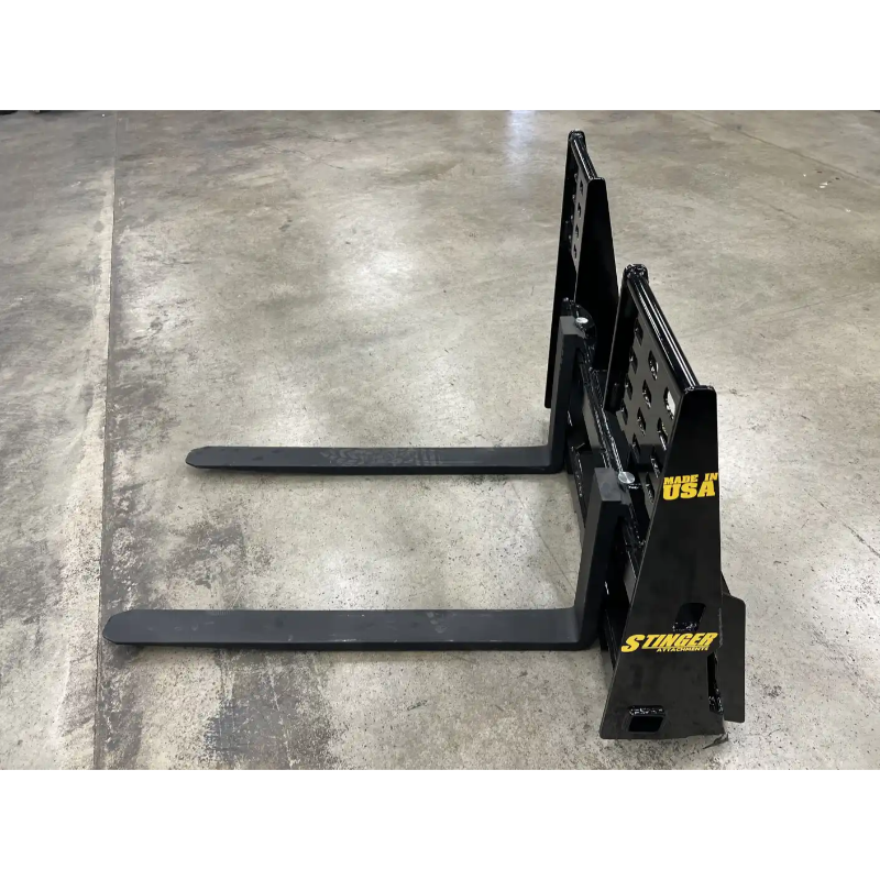 Black painted steel pallet fork attachment with two long horizontal tines and reinforced vertical mounting plate on concrete floor