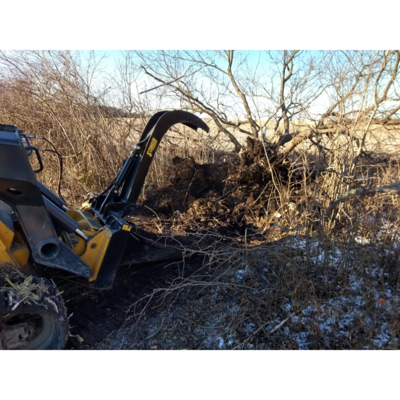 Yellow skid steer with black curved stump grapple attachment holding uprooted tree and branches on dirt terrain