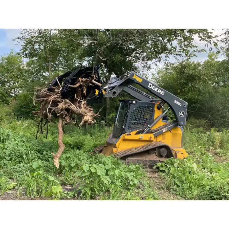Yellow Deere skid steer with black clam grapple attachment lifting large root ball in overgrown field area