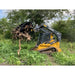 Yellow compact track loader with black clam grapple attachment lifting large uprooted tree with roots in green field