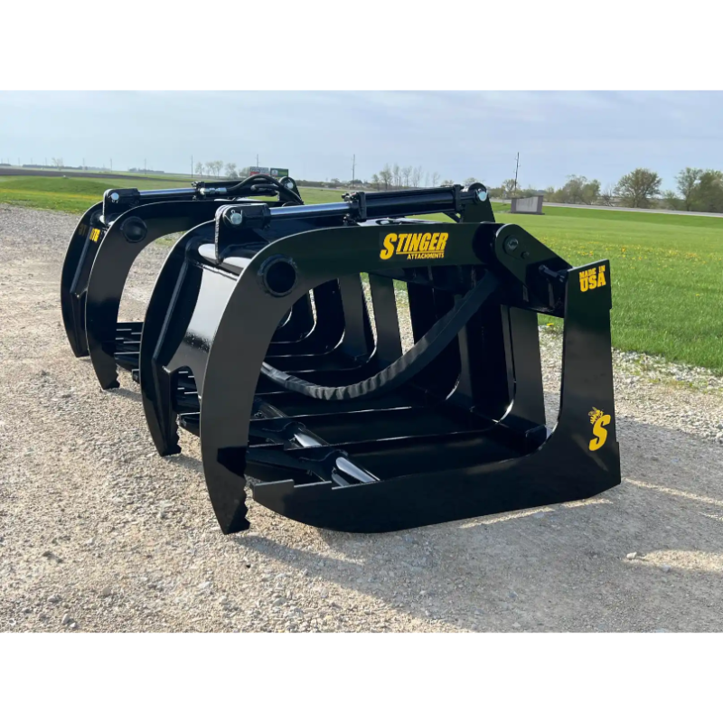 Skid steer root grapple attachment with thick curved tines, hydraulic arms, and black painted steel frame on gravel road