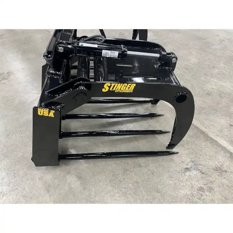 Black steel grapple attachment with four sharp tines and hydraulic arm on a concrete floor indoors