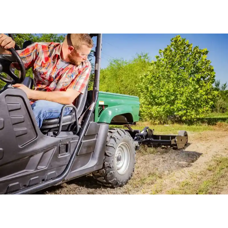 Operator in plaid shirt driving green utility vehicle towing Swisher 48" Driveway Grader on dirt surface, gallery position 1