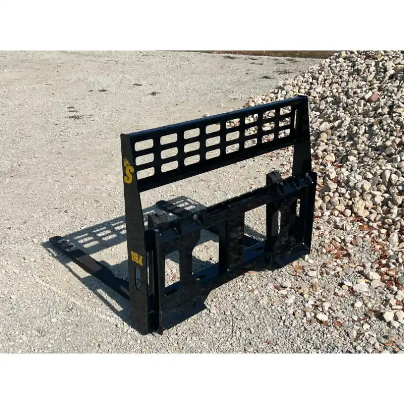 Black steel skid steer pallet fork mounting frame with open grid backrest on gravel surface near stone pile