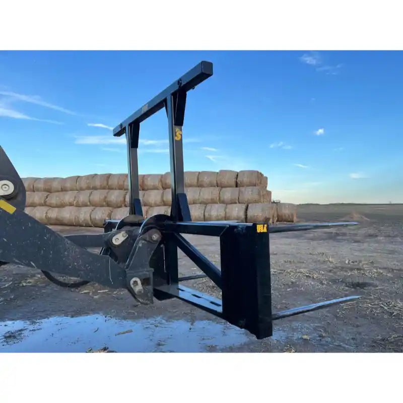 Black steel bale spear skid steer attachment with dual vertical support bars and dual horizontal tines in outdoor dirt setting