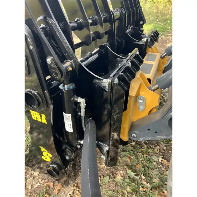 Close-up of black steel clam grapple attachment with hydraulic hoses and yellow skid steer mounting bracket outdoors on dirt and leaves