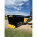 Black steel skid steer snow pusher attachment with yellow bolt-on cutting edge on gravel and grass under cloudy sky